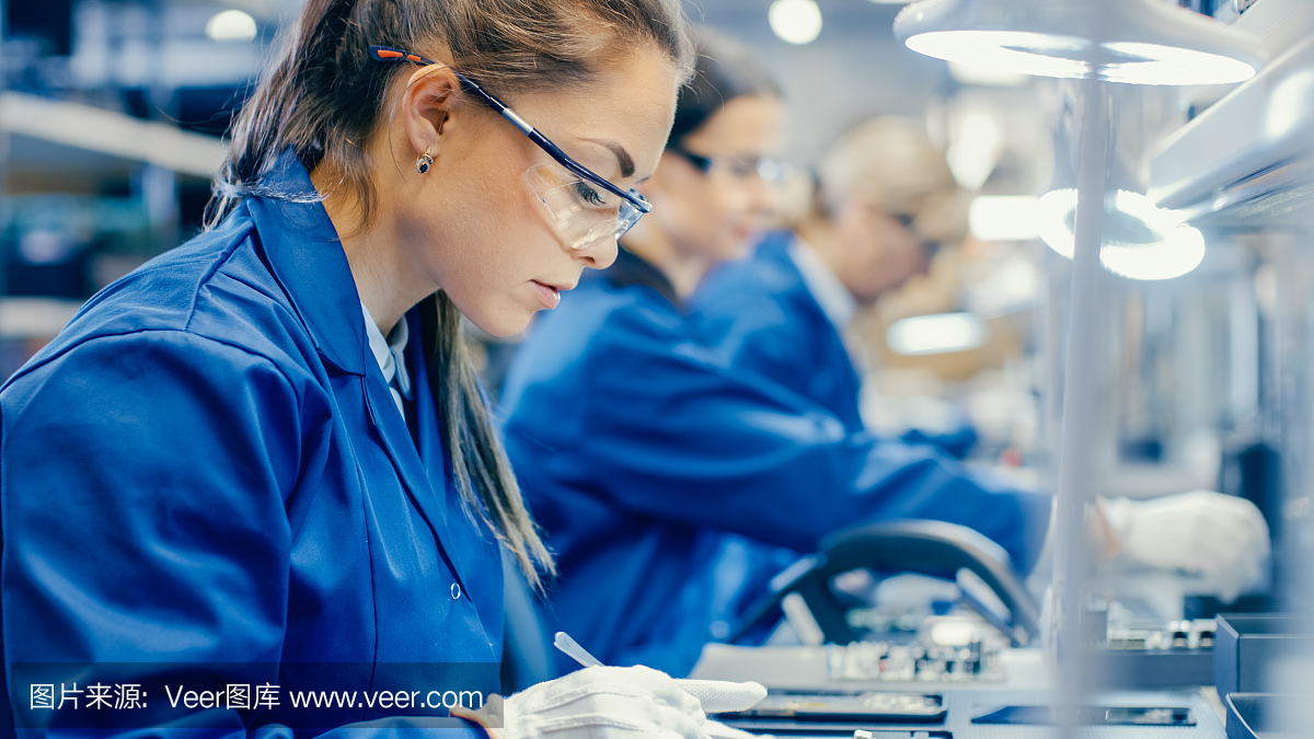 穿着蓝色工作服和防护眼镜的电子厂女工正在用镊子组装智能手机的印刷电路板。拥有更多员工的高科技工厂。